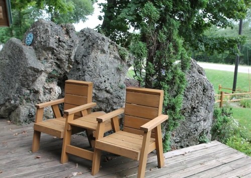 Two wooden chairs on a deck beside large rocks and greenery, with a grassy area and road in the background.