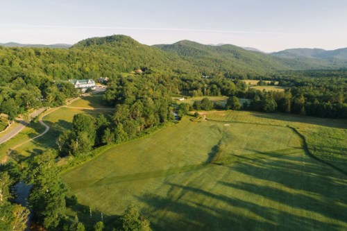 Aerial view of a lush green landscape with rolling hills, a golf course, and a distant building surrounded by trees.