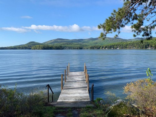 A wooden dock extends into a calm lake, surrounded by green hills and a clear blue sky.