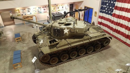 A military tank displayed indoors, featuring a star emblem, with an American flag in the background.