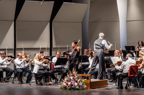 A conductor leads an orchestra on stage, with musicians playing string instruments and a floral arrangement in front.