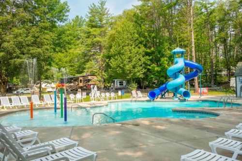 A swimming pool with a blue water slide, surrounded by lounge chairs and trees in a campground setting.