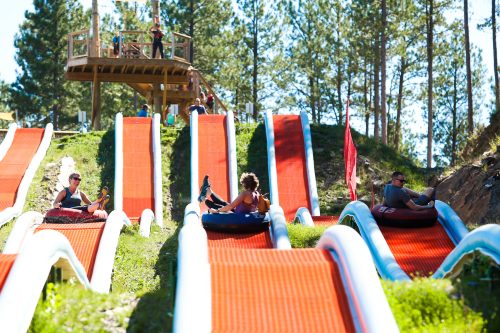 Two people slide down colorful, winding water slides in a forested area, with a wooden platform in the background.