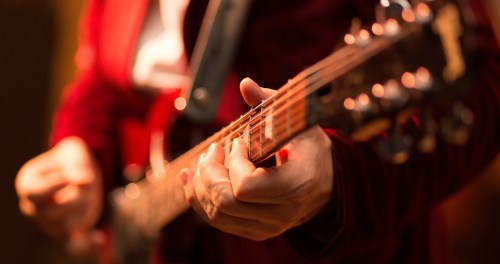 A close-up of a musician's hands playing a red electric guitar on stage.