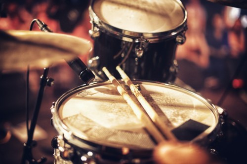 Close-up of a drum set with drumsticks resting on the snare drum, blurred audience in the background.