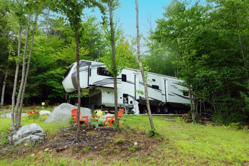A large RV parked in a wooded area, surrounded by greenery and a small seating area with red chairs.