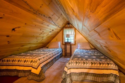 Cozy attic bedroom with two twin beds, wooden walls, and a small table by a window. Warm, inviting atmosphere.