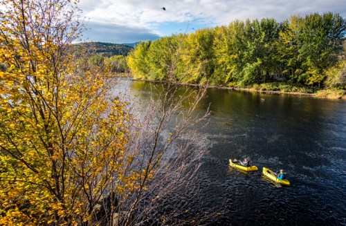 Two kayakers paddle on a calm river surrounded by vibrant autumn foliage and distant hills under a partly cloudy sky.