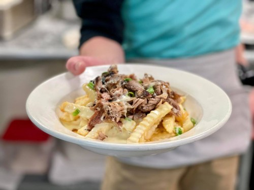 A person holds a plate of pasta topped with pulled pork and green onions in a restaurant setting.