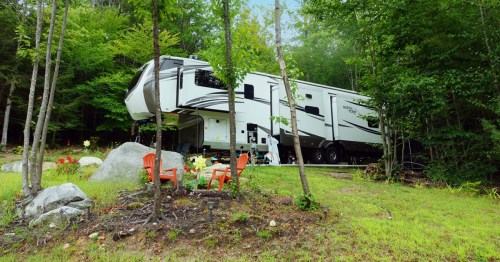 A large RV parked in a wooded area, surrounded by greenery and two red chairs on a grassy patch.