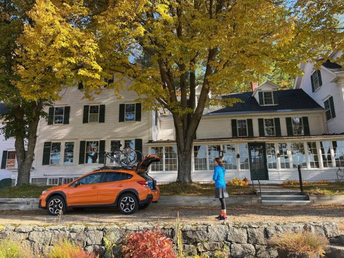 A person in a blue jacket stands near an orange car with a bike rack, in front of a large white house surrounded by autumn trees.