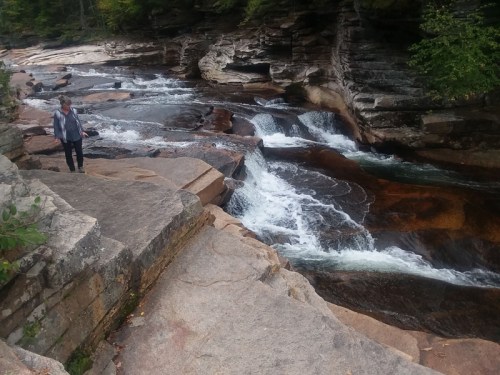 A person stands near a rocky riverbank with flowing water and lush greenery in the background.