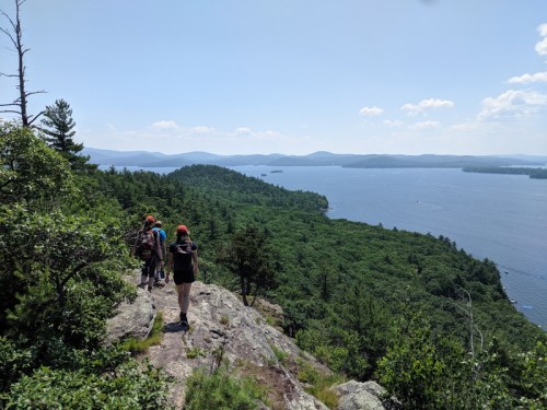 Hikers on a rocky trail overlooking a lush green landscape and a lake under a clear blue sky.