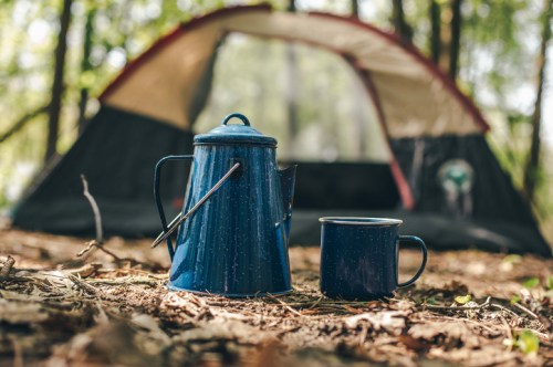 A blue camping kettle and mug sit on the forest floor in front of a tent, surrounded by leaves and trees.