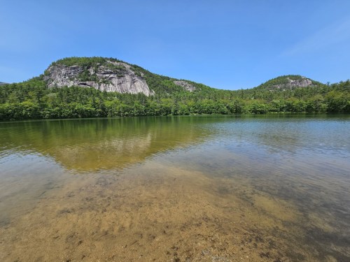 A serene lake surrounded by lush green hills under a clear blue sky, with reflections on the water's surface.