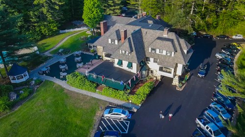 Aerial view of a large, charming house surrounded by trees, with a parking lot and outdoor seating area.