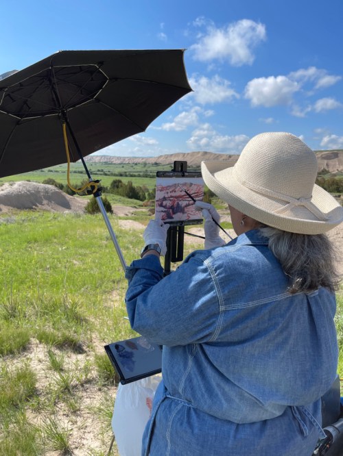 An artist in a sun hat paints outdoors under an umbrella, surrounded by green hills and blue skies.