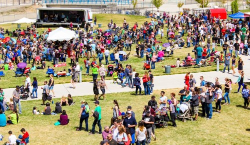 A large crowd gathers at an outdoor event with tents, food stalls, and activities on a sunny day.