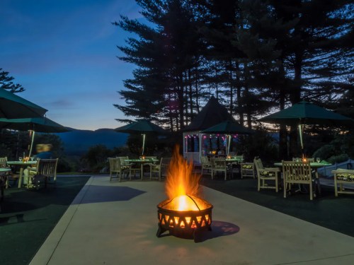 A cozy outdoor setting at dusk with a fire pit, tables, and umbrellas surrounded by trees and mountains in the background.