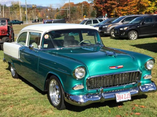 A classic teal and white 1955 Chevrolet parked on grass, surrounded by trees with autumn foliage.