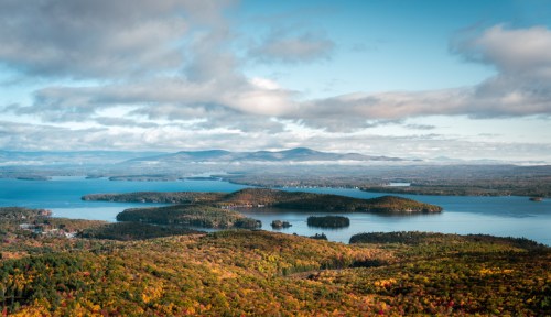 A scenic view of a lake surrounded by colorful autumn foliage and distant mountains under a partly cloudy sky.