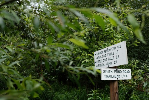 Trail sign among greenery, indicating distances to Shaker Reservoir, Big Dog Falls, Little Dog Falls, and Smith Pond Road.