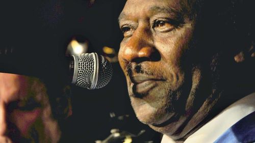 A close-up of a man singing into a microphone, with a soft focus on a musician in the background.