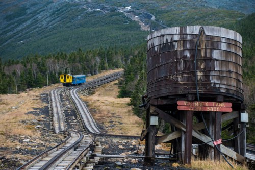 A weathered water tank beside a winding railway track with a blue and yellow train in a mountainous landscape.