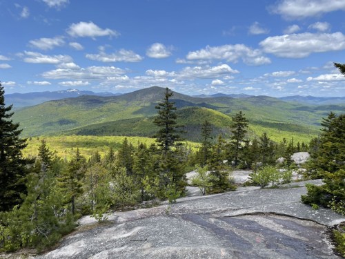 A scenic view from a rocky summit, showcasing lush green mountains and a blue sky with fluffy clouds.