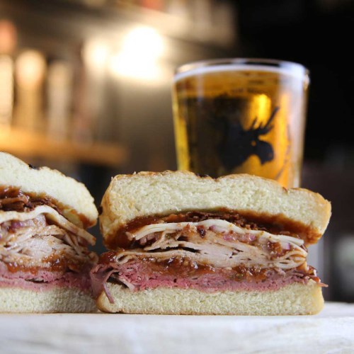 A close-up of a sliced sandwich filled with meats and sauce, alongside a glass of beer in the background.