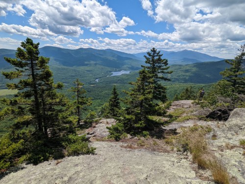 A scenic view from a rocky overlook, featuring lush green mountains and a river under a partly cloudy sky.