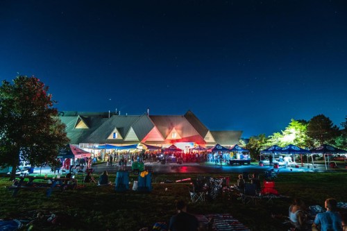 A lively outdoor event at night, with people gathered under tents and colorful lights illuminating a building.