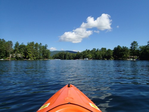 A view from an orange kayak on calm water, surrounded by trees and a clear blue sky with a few clouds.