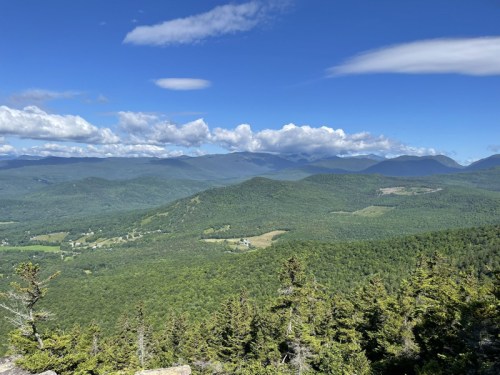A panoramic view of lush green mountains and valleys under a bright blue sky with scattered clouds.