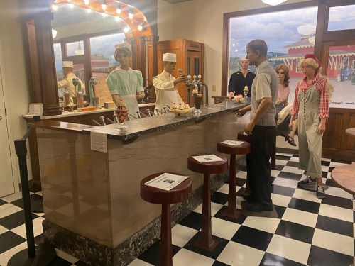 A vintage diner scene with mannequins at a counter, featuring ice cream sundaes and a checkered floor.