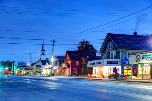 A quiet street at dusk featuring shops, a church with a steeple, and a house with solar panels.