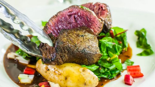 A close-up of a gourmet dish featuring tender beef, greens, and a potato, garnished with sauce.