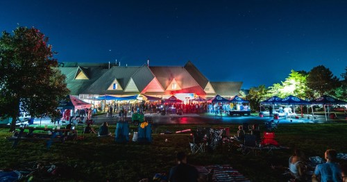 A lively outdoor event at night with people gathered near a brightly lit building and tents under a starry sky.