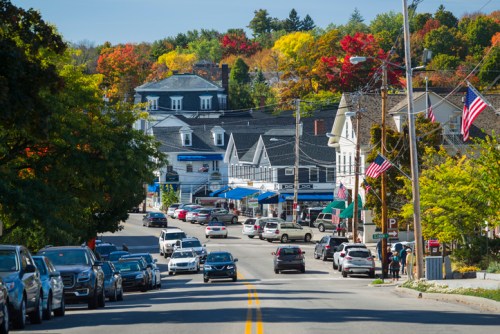 A scenic street view of a small town with colorful autumn trees, shops, and American flags lining the road.