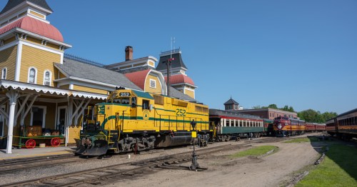 A vintage yellow and green train at a historic station, with passenger cars and a clear blue sky in the background.