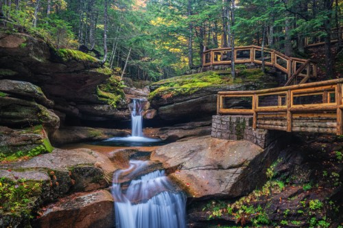 A serene forest scene featuring a waterfall cascading over rocks, with wooden walkways and lush greenery surrounding it.