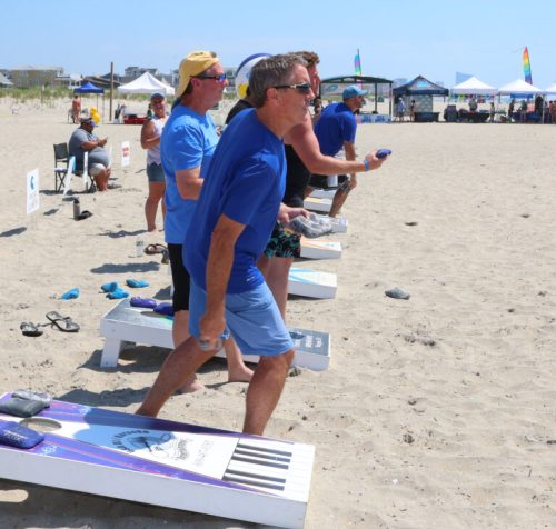 Three men in blue shirts play cornhole on a sandy beach, with tents and people in the background.