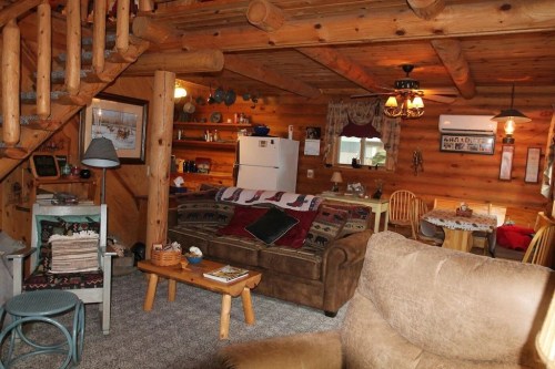 Cozy log cabin interior with a living area, kitchen, and dining space, featuring wooden decor and warm lighting.