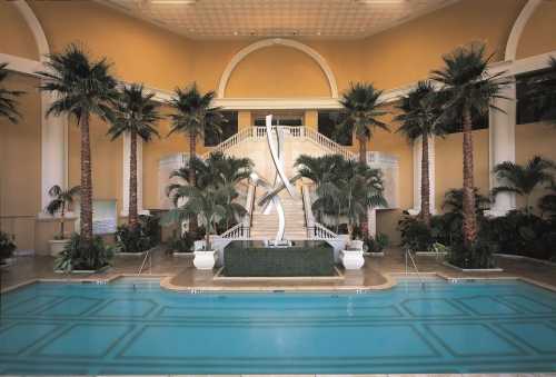 Luxurious indoor pool area with palm trees, a grand staircase, and a modern sculpture in a serene setting.