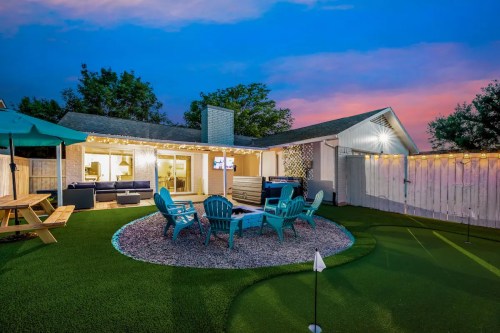A cozy backyard with teal chairs around a fire pit, a picnic table, and a putting green, illuminated by string lights at dusk.