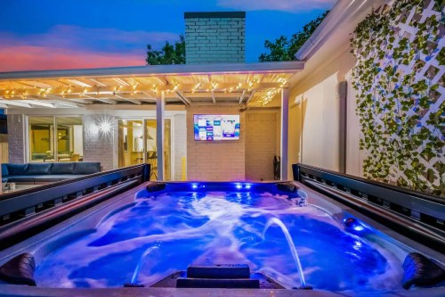 A hot tub with blue lighting, surrounded by a patio and a TV, under a twilight sky with string lights above.