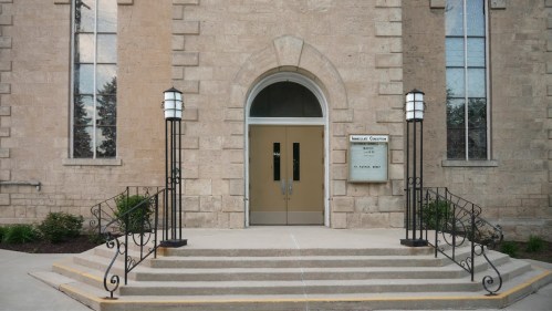 Entrance of a stone building with a large arched door, flanked by lanterns and steps leading up to it.