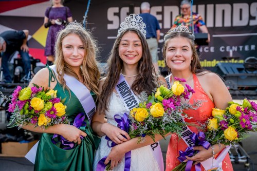 Three young women in formal dresses hold bouquets, smiling and wearing tiaras at an outdoor event.