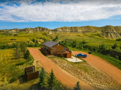 Aerial view of a brown house on a dirt road, surrounded by green fields and hills under a blue sky.