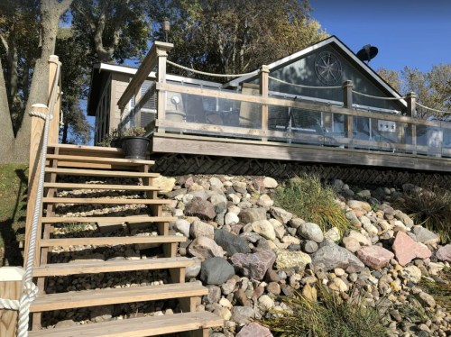 A wooden staircase leads up to a house with a deck, surrounded by rocks and greenery on a sunny day.